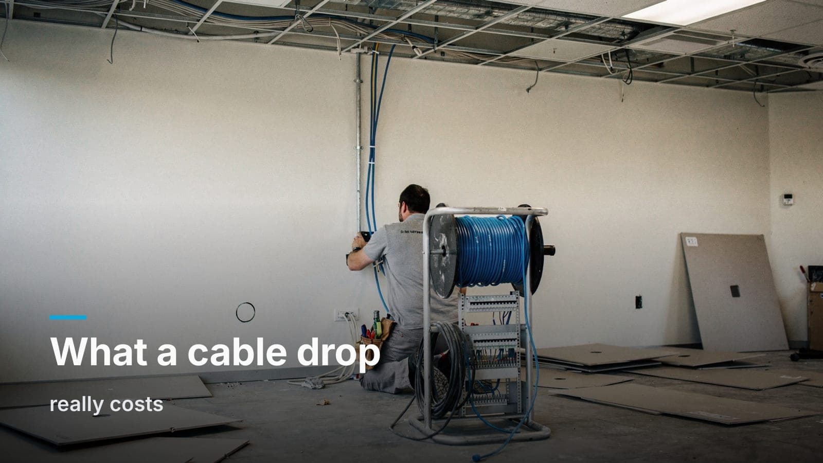 Installer kneeling at a low wall plate in an empty office, blue Cat6 spool on a caddy, cable runs into open ceiling grid.
