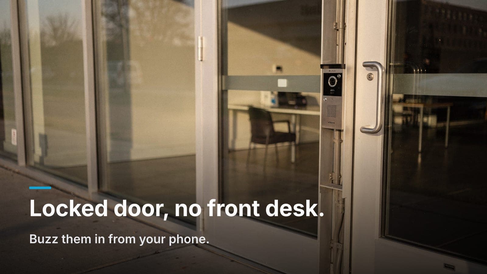 Video intercom station mounted on the frame of a locked commercial glass storefront door in afternoon sun.