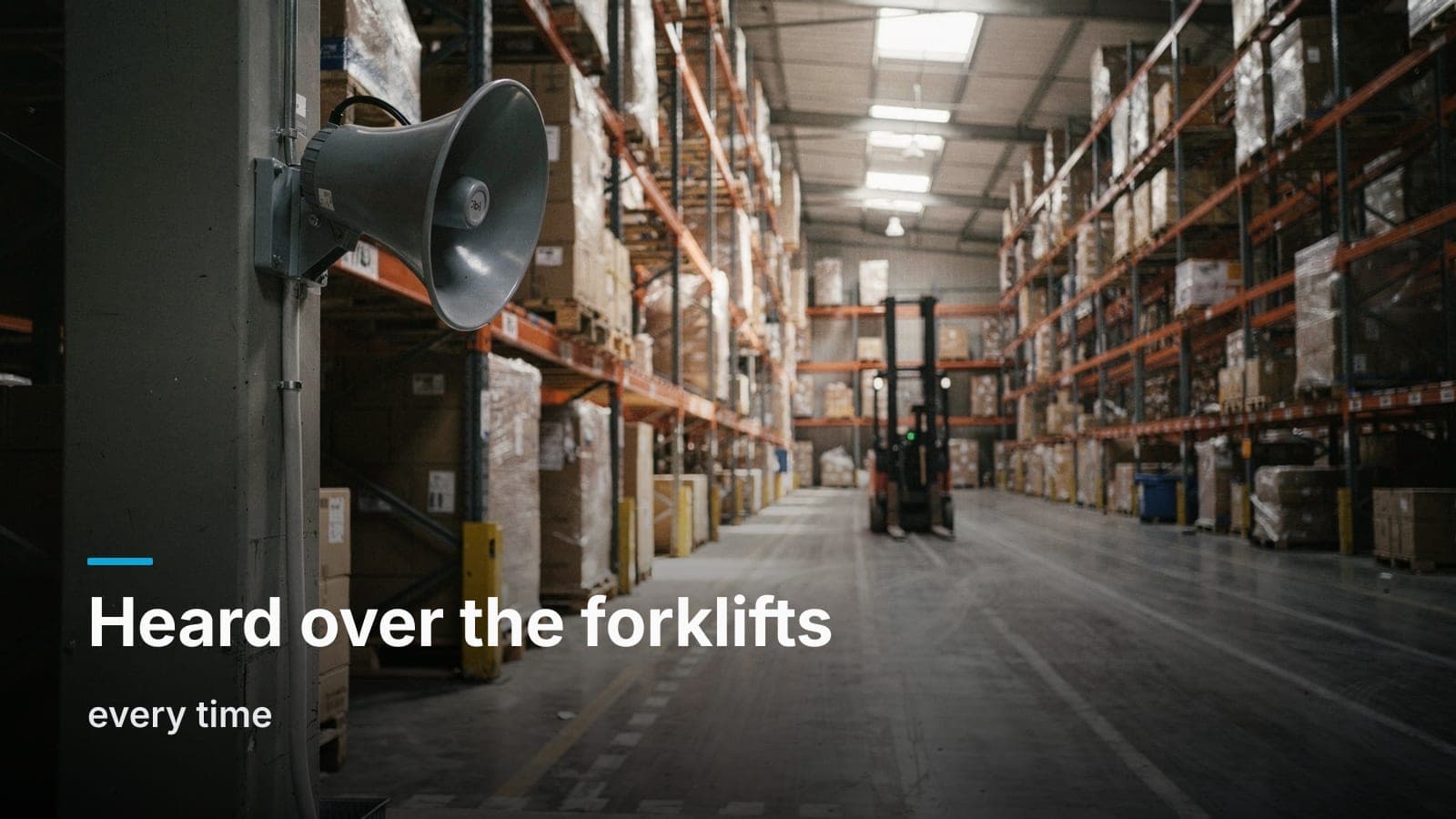 A gray horn loudspeaker mounted high on a steel column in a warehouse aisle, with a parked forklift down the row and daylight from skylights overhead.