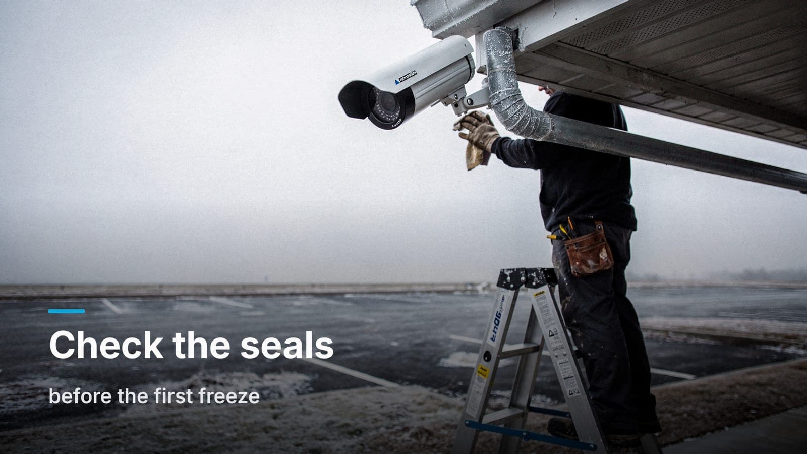 A technician on a ladder wipes frost from an outdoor security camera lens under a building eave on a gray winter morning.