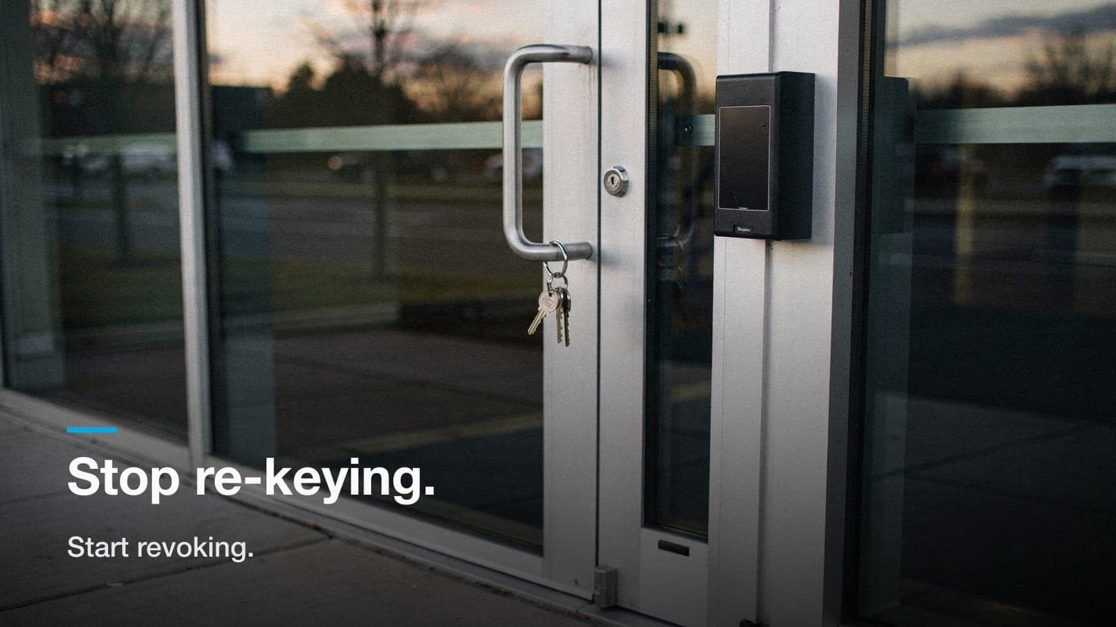 Keys hanging from the lock of a clean glass office door beside a newly mounted badge reader.