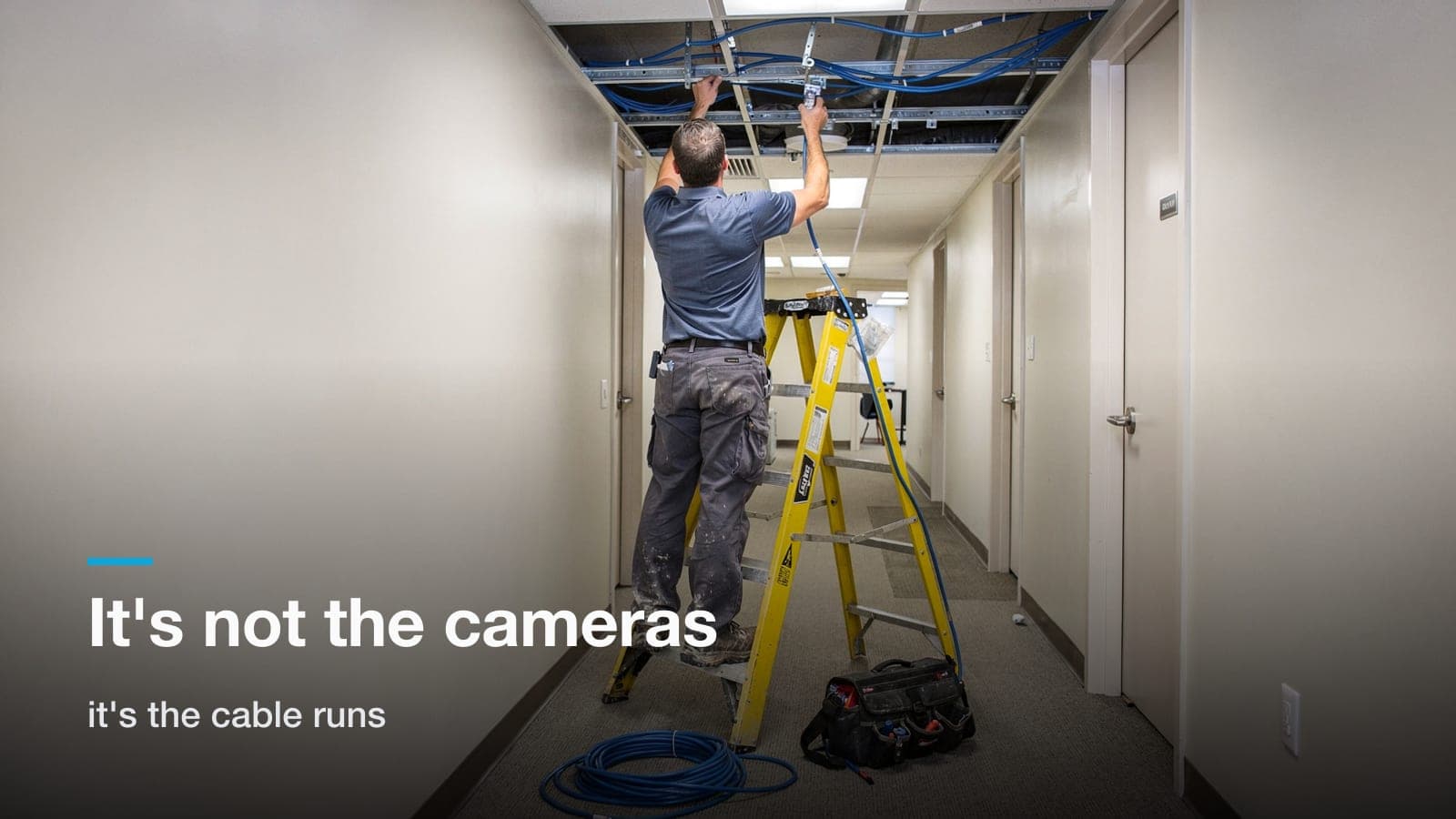 Installer on a ladder in an office hallway feeding Cat6 cable above an open drop-ceiling tile.