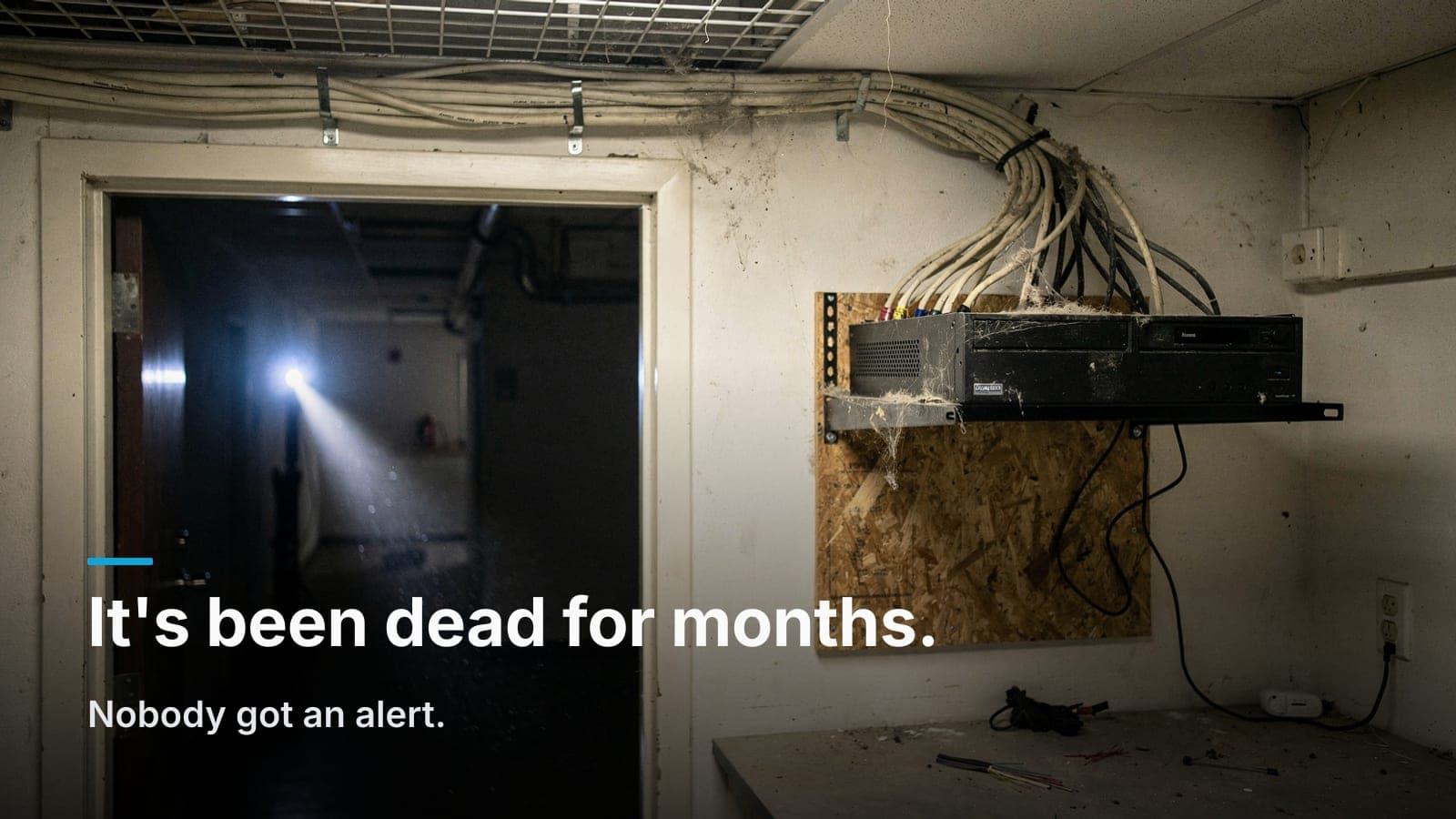 Dusty network closet with a wall-mounted camera recorder, dark drive light, cobwebs on cables, flashlight beam.