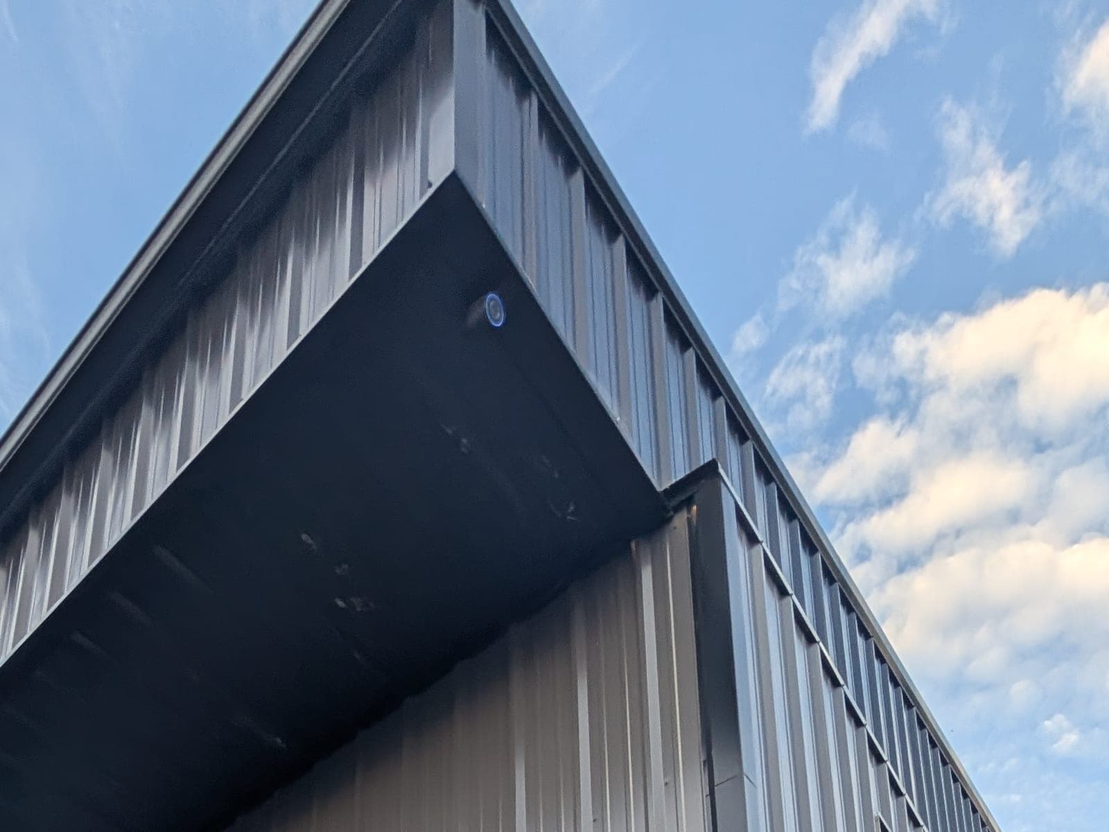 Exterior security camera mounted under the eave at the corner of a black metal commercial building